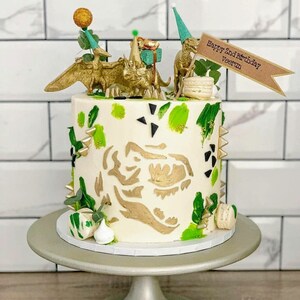 May include: A white birthday cake decorated with gold dinosaur figurines, green and gold brushstrokes, and a gold dinosaur head silhouette. The cake is topped with a banner that reads "Happy 2nd Birthday, Veerun."