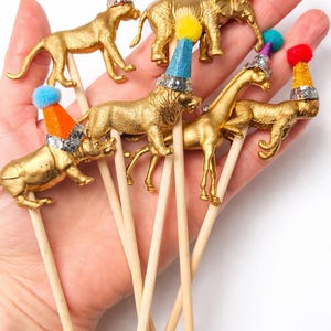 以下が含まれることがあります： 金色の動物の置物。ライオンやゾウなど、カラフルなパーティーハットとポンポンが付いています。木製の棒に取り付けられており、ケーキトッパーやパーティーの装飾に最適です。帽子は様々な色があります。