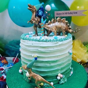 May include: A two-tiered birthday cake with green and white frosting, decorated with gold dinosaur figurines and colourful candies. The cake is topped with a banner that reads "Happy 1st Birthday Rex". Balloons in shades of blue and yellow are in the background.