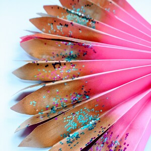 May include: A pink and gold paper fan with glitter. The fan is folded and has a gradient of colour from pink to gold. The glitter is a mix of teal, pink, and gold.