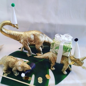 May include: Three gold dinosaur ornaments, including a Brachiosaurus and a Triceratops, are displayed on a large green leaf. One Triceratops wears a party hat and another has a gift box on its back. Gold confetti is scattered around.