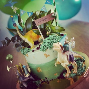 May include: A two-tiered birthday cake decorated with green frosting, gold leaf accents, and toy dinosaurs. The cake is adorned with greenery, a banner that reads "FREDDI", and a sign that says "1 Today!" Green and teal balloons are in the background.