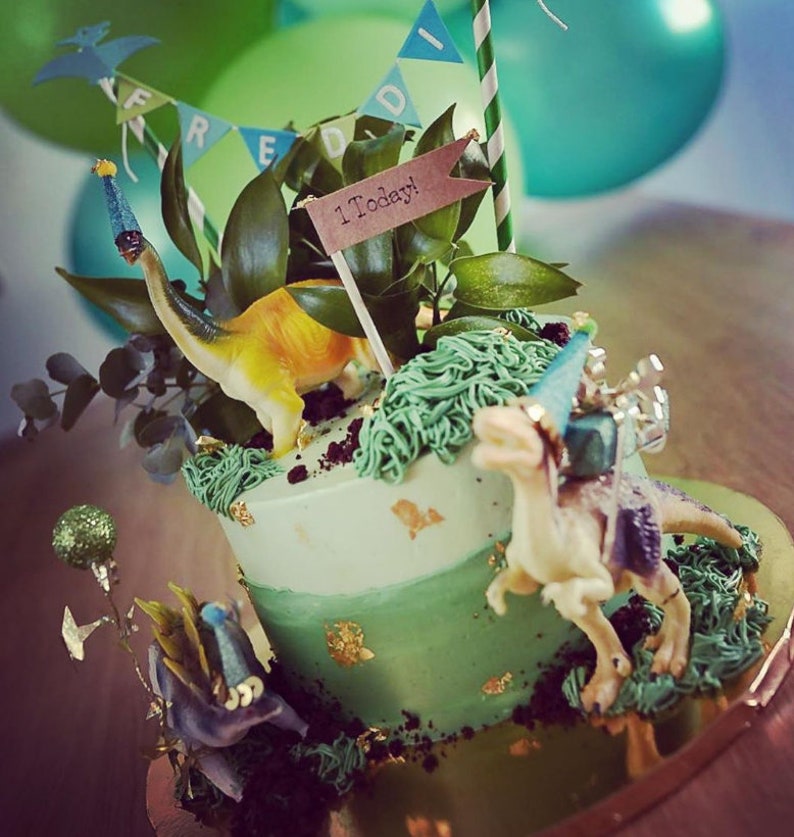May include: A two-tiered birthday cake decorated with green frosting, gold leaf accents, and toy dinosaurs. The cake is adorned with greenery, a banner that reads "FREDDI", and a sign that says "1 Today!" Green and teal balloons are in the background.