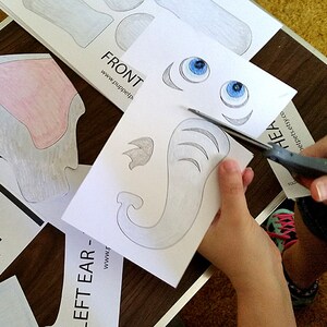 Puede incluir: Un ni&ntilde;o corta una plantilla de elefante de papel con tijeras. La plantilla incluye la cabeza, las orejas y la trompa del elefante. El papel es blanco con marcas de l&aacute;piz gris. El texto en el papel dice "LEFT EAR - www.puppetp.com".