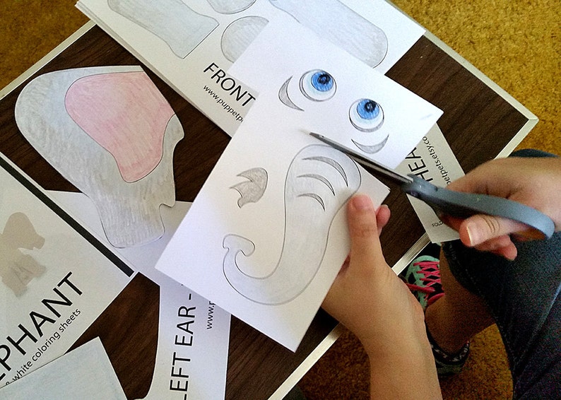 Puede incluir: Un ni&ntilde;o corta una plantilla de elefante de papel con tijeras. La plantilla incluye la cabeza, las orejas y la trompa del elefante. El papel es blanco con marcas de l&aacute;piz gris. El texto en el papel dice "LEFT EAR - www.puppetp.com".