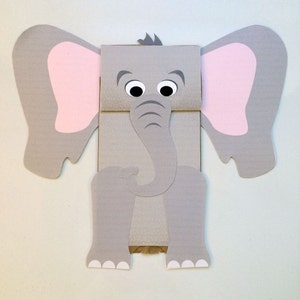 Puede incluir: Una manualidad con forma de elefante gris hecha de cartón con el interior de las orejas rosa. El elefante tiene ojos grandes y redondos, una larga trompa y orejas grandes. Las patas y los pies también son grises con detalles blancos. La manualidad está diseñada para estar de pie.