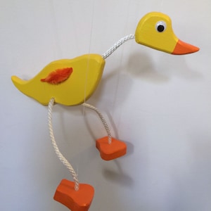 May include: A yellow wooden puppet duck with orange feet and a red and orange feather on its back. The duck is suspended by strings attached to its head, wings, and feet.