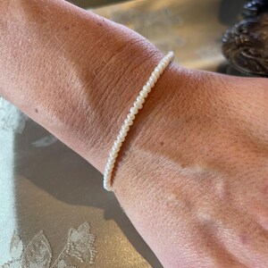 Könnte beinhalten: Ein zartes Armband aus kleinen, runden, weißen Perlen. Das Armband ist aufgefädelt und bildet einen durchgehenden Strang aus glänzenden Perlen. Der Schmuck wird am Handgelenk getragen und zeigt die elegante Schlichtheit des Designs.