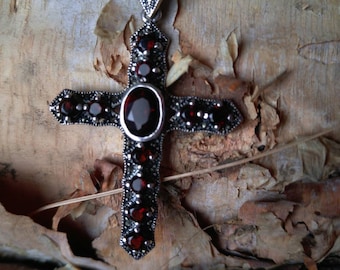 May include: A silver cross pendant with red gemstones set in a marquetry design. The pendant is on a wooden surface.