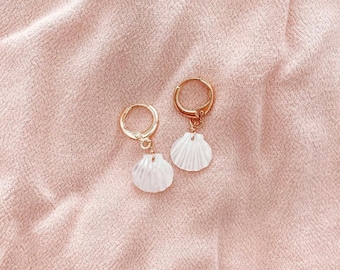 May include: A pair of gold-colored hoop earrings with white seashell charms. The earrings are displayed on a pink fabric background. The seashell charms have a detailed, textured surface.
