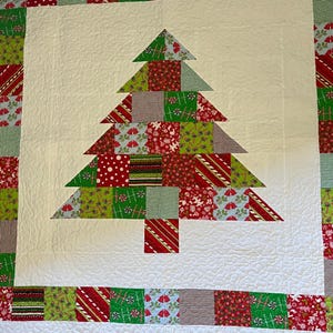 May include: A white quilt with a patchwork Christmas tree design. The tree is made of green, red, and white fabric squares. The quilt is bordered with a red and white striped fabric.