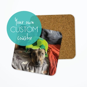 May include: A brown and black dog wearing a green and white football hat is lying on a grey and red blanket. The dog is looking at the camera. The image is on a cork coaster.