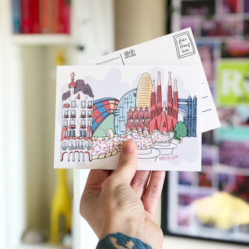 May include: A colorful postcard featuring a cartoon illustration of the Barcelona skyline. The postcard includes iconic landmarks like the Sagrada Familia, Park G&uuml;ell, and Casa Batll&oacute;. The text "Barcelona" is printed at the bottom of the postcard.