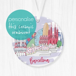 May include: A white ceramic ornament with a colorful illustration of the Barcelona skyline, including the Sagrada Familia, Park Güell, and other iconic landmarks. The ornament is round and has a white ribbon attached for hanging. The text "Barcelona" is written in pink at the bottom of the ornament.