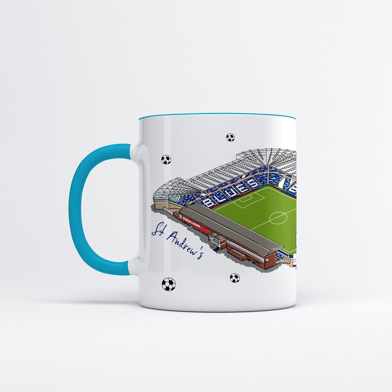 May include: White ceramic mug with a blue rim and handle. The mug features a detailed illustration of St. Andrew's Stadium, home of Birmingham City Football Club. The stadium is depicted in a bird's-eye view, with the words "Blues" visible on the pitch. The text "St. Andrew's" is written in cursive below the stadium.