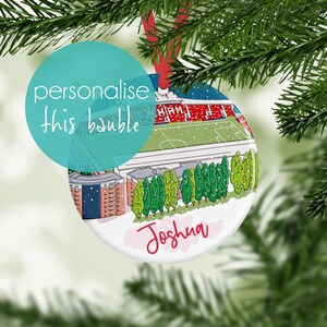 May include: A white, round Christmas ornament with a stadium illustration. The ornament features a red ribbon for hanging and the name "Joshua" in red script. The text "personalise this bauble" is also present.