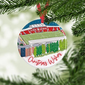 May include: A round Christmas ornament featuring a stadium scene with the text "Wrexham" and "Christmas Wishes" in red script. The ornament has a red ribbon for hanging and is set against a backdrop of evergreen branches.