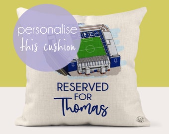 May include: A white cushion with a blue and white illustration of a football stadium. The text "RESERVED FOR Thomas" is printed on the cushion.