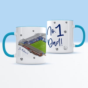 May include: White ceramic mug with a blue rim and handle. One side features a cartoon illustration of St. Andrew's Stadium, home of Birmingham City Football Club. The other side has the text "No. 1 Dad!" in blue cursive writing.
