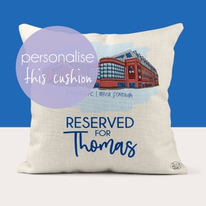 May include: A white cushion with a blue and red illustration of a stadium. The text "RESERVED FOR Thomas" is printed in blue on the cushion.