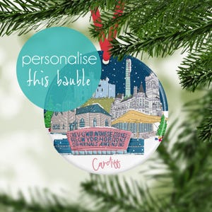 May include: A round Christmas bauble featuring a winter scene of Cardiff, Wales, with buildings and text. The bauble has a red ribbon for hanging and the words "personalise this bauble" are on a teal circle.