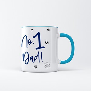 May include: White ceramic mug with a blue rim and handle. The mug has a blue inscription that reads "No. 1 Dad!" with soccer ball graphics.
