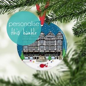 May include: A round ceramic Christmas ornament featuring a dark blue winter scene with a black and white timber-framed building, snow-covered ground, green pine trees, and a small figure in a red coat carrying a sack, suspended by a red ribbon.