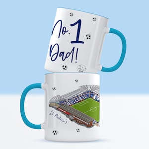 May include: Two white ceramic mugs with blue handles. The top mug has the text "No. 1 Dad!" in blue lettering. The bottom mug has a graphic of a soccer stadium with the text "St. Andrew's" in blue lettering.