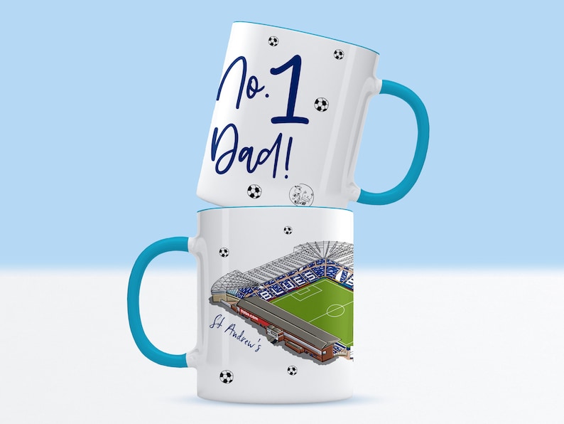 May include: Two white ceramic mugs with blue handles. The top mug has the text "No. 1 Dad!" in blue lettering. The bottom mug has a graphic of a soccer stadium with the text "St. Andrew's" in blue lettering.