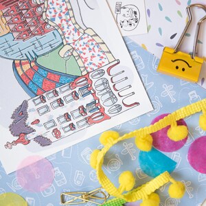 May include: A white postcard with a colorful illustration of a building with a red, blue, and green design. The postcard is surrounded by yellow pom-poms, a blue and white patterned paper, and a yellow paperclip with a smiley face.