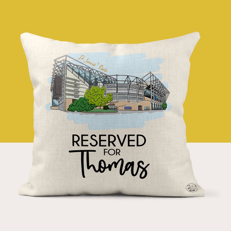 May include: A white cushion featuring a black and white illustration of St. James' Park, a football stadium. The cushion has the text "RESERVED FOR Thomas" printed on it.