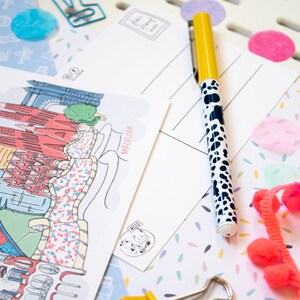 May include: A white postcard with a colorful illustration of the Barcelona skyline. The postcard has the text "Barcelona" written on it. A yellow pen with black polka dots is lying on top of the postcard.