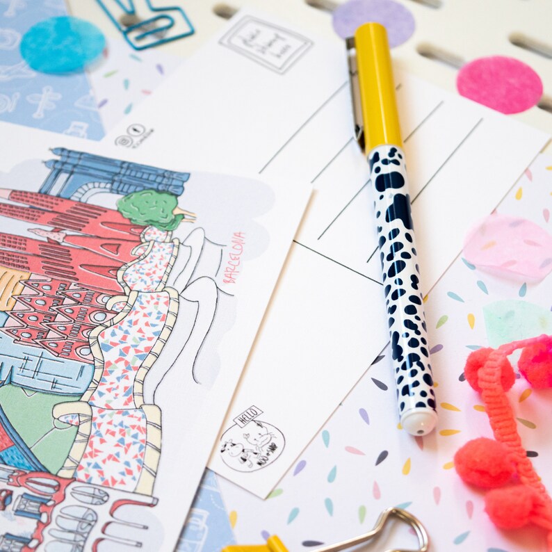May include: A white postcard with a colorful illustration of the Barcelona skyline. The postcard has the text "Barcelona" written on it. A yellow pen with black polka dots is lying on top of the postcard.
