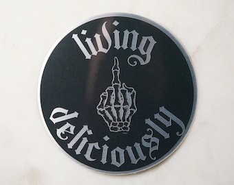 May include: Black and silver round sticker with the text "living deliciously" in a gothic font. A skeleton hand giving the middle finger is in the center of the sticker.