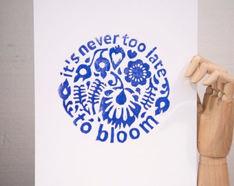 Puede incluir: Impresión artística blanca con un diseño floral azul en forma circular. El texto "it's never too late to bloom" está impreso en azul. La impresión está sujeta por una mano de madera.