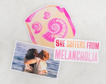May include: Three stickers on a white surface. One sticker features a pink and beige seashell design. Another shows a photo of two people in the water. The third sticker reads "She Suffers From Melancholia."
