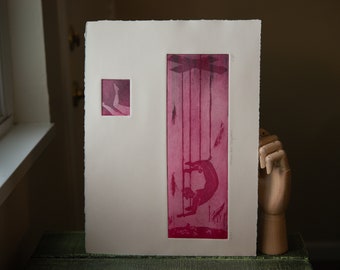 Puede incluir: Una impresión rosa y blanca de una persona colgada de cuerdas, con una impresión más pequeña de las piernas de una persona en la esquina superior izquierda. La impresión está enmarcada y sobre una superficie de madera.