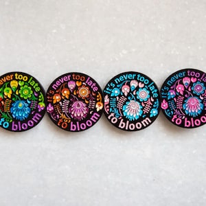 May include: Four round black enamel pins with colorful floral designs and the text "It's never too late to bloom".
