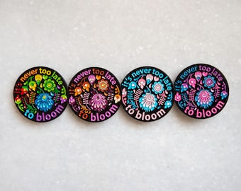 Puede incluir: Cuatro pines redondos de esmalte negro con diseños florales coloridos y el texto "Nunca es demasiado tarde para florecer".