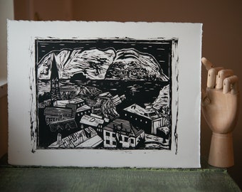 Puede incluir: Grabado en linóleo en blanco y negro de una ciudad con montañas en el fondo. La impresión está enmarcada con un borde negro.