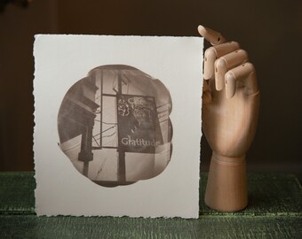 Puede incluir: Una fotografía en sepia de un edificio con un letrero que dice "Gratitude" en un marco circular. La fotografía está montada en una cartulina blanca con un borde festoneado.