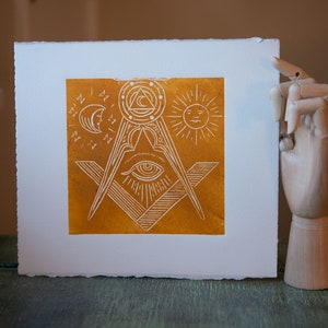 May include: A white print on a yellow background with a compass and square, an eye, a sun, and a moon. The print is framed in a white mat.