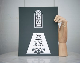 Puede incluir: Una impresión en blanco y negro con el texto "girls like girls who like girls" en una fuente gótica. La impresión está sobre un fondo gris oscuro y presenta un diseño blanco de una ventana de iglesia con un arco apuntado.