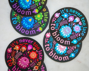 May include: Four round black and white stickers with colorful floral designs and the text "it's never too late to bloom" repeated around the edge of each sticker.