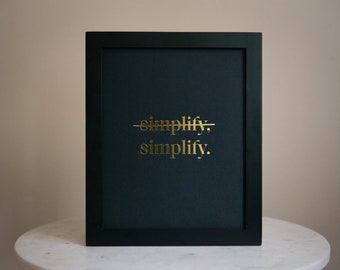 Puede incluir: Impresión artística enmarcada en negro con el texto dorado "simplify. simplify." sobre un fondo verde oscuro. La palabra "simplify" está tachada. El arte se exhibe sobre una superficie de mármol blanco.