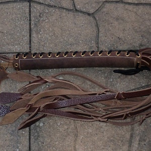 May include: A brown leather whip with a braided handle and multiple leather strands. The handle is laced with black cord and has gold-colored eyelets. A small leather tag is attached to the handle.