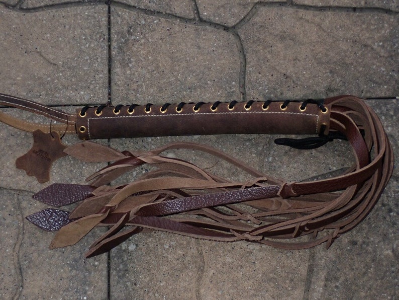 May include: A brown leather whip with a braided handle and multiple leather strands. The handle is laced with black cord and has gold-colored eyelets. A small leather tag is attached to the handle.