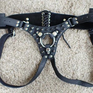 May include: Black leather harness with silver-toned studs and a large metal ring. The harness features adjustable straps and a lace-up detail in the back. The design includes a waist belt and suspender-style straps.