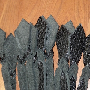 May include: Close-up of several black leather strips, some with a textured surface, arranged in a braided pattern. The strips are knotted at intervals, creating a decorative effect. The background is a light brown wooden surface.