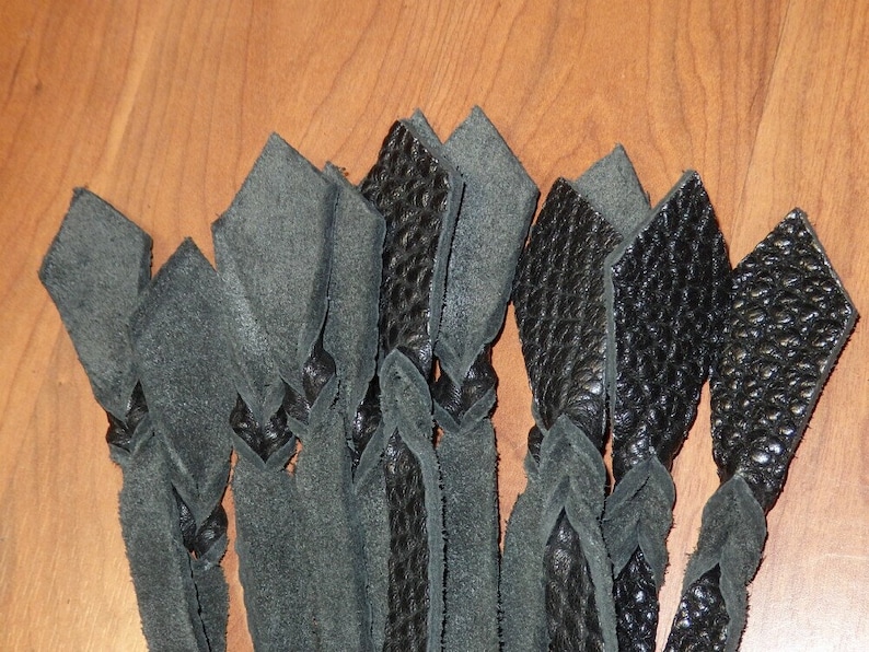 May include: Close-up of several black leather strips, some with a textured surface, arranged in a braided pattern. The strips are knotted at intervals, creating a decorative effect. The background is a light brown wooden surface.